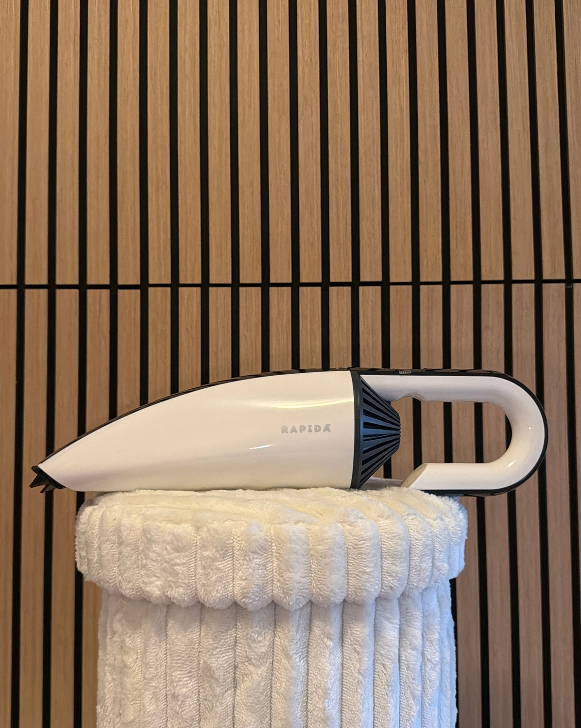 Erfolg – ​​​​Verkauf von HYLA-Produkten – Weißer Handstaubsauger mit der Aufschrift RAPIDA, der auf einer weißen, plüschigen, strukturierten Oberfläche mit einem vertikalen Holzlamellenhintergrund ruht