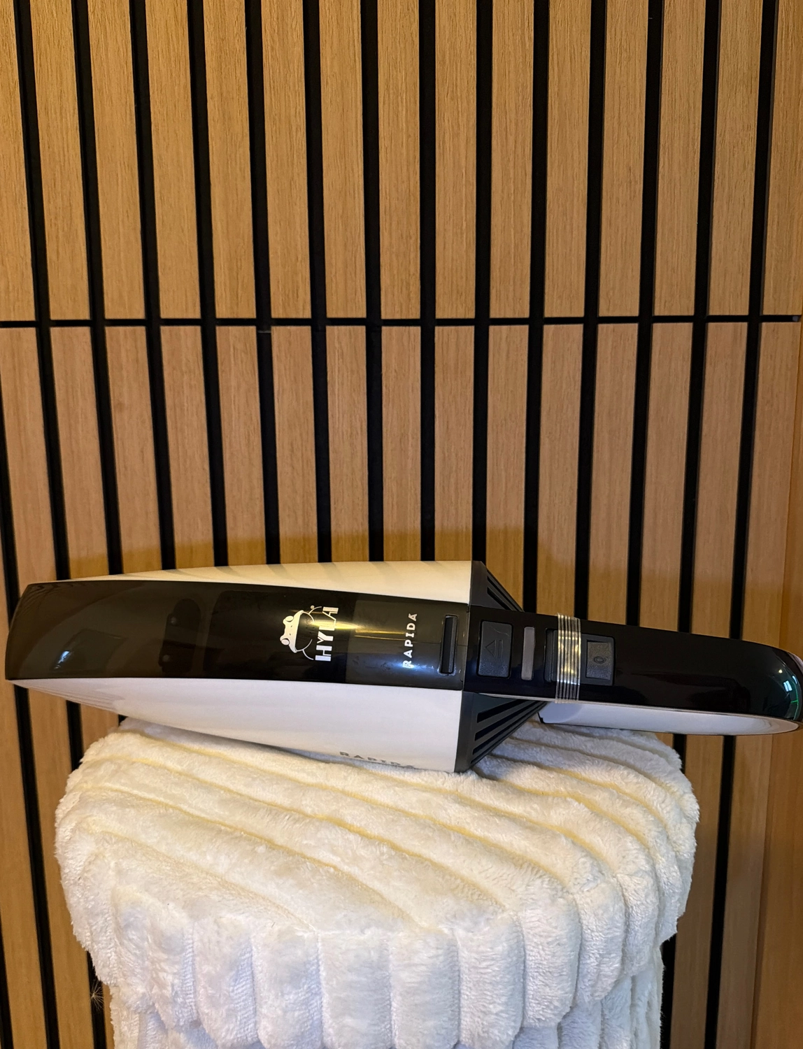 Erfolg – ​​​​Verkauf von HYLA-Produkten – Schwarz-weißer Handstaubsauger mit der Marke „HYA RAPIDA“, der auf einem weichen, weißen, strukturierten Handtuch vor einem Hintergrund aus Holzlatten ruht