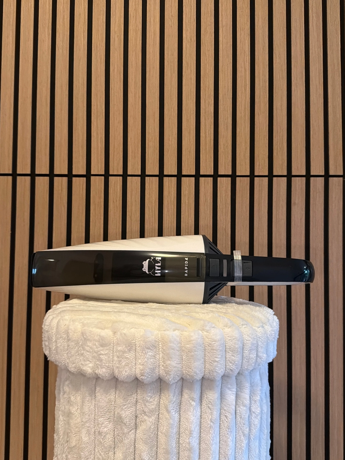 Erfolg – Verkauf von HYLA-Produkten – Schwarz-weißer Handstaubsauger Hyla Rapida, der auf einem weißen strukturierten Hocker an einer Holzlattenwand ruht