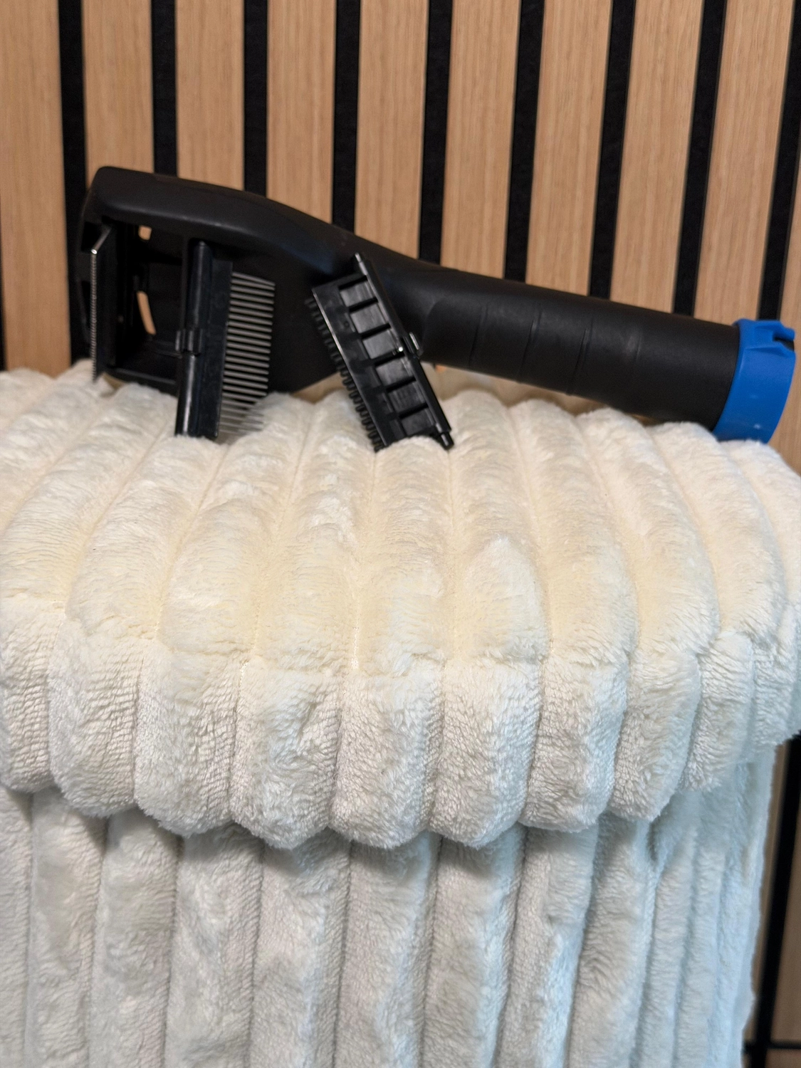 Erfolg – Verkauf von HYLA-Produkten – Schwarzes Haustierpflegegerät mit abnehmbaren Kammaufsätzen, das auf einer plüschigen weißen Decke vor einem hölzern und schwarz gestreiften Hintergrund ruht.